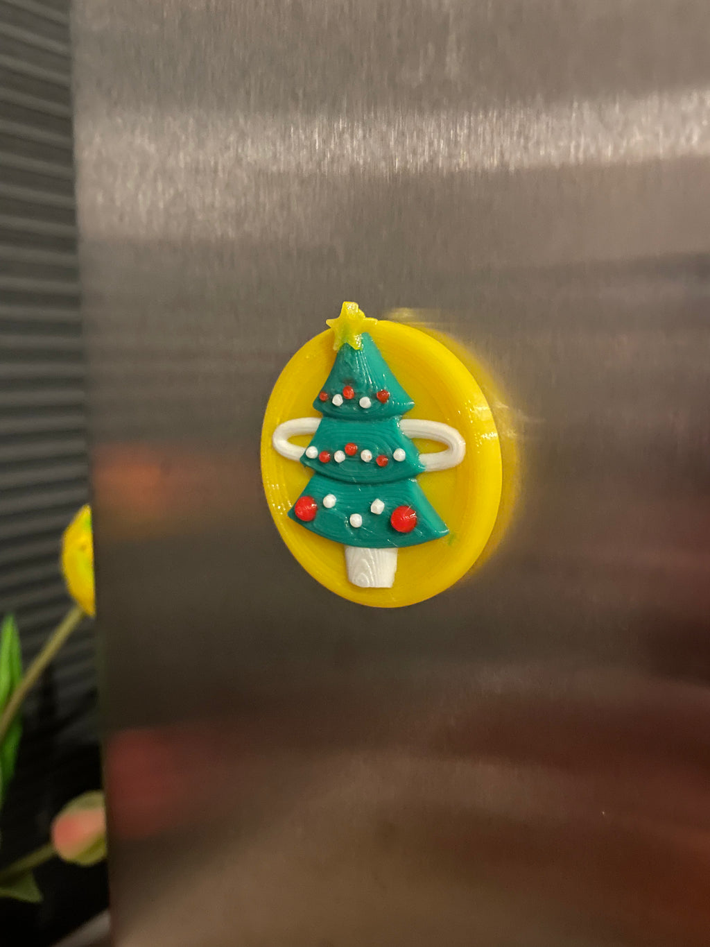 3D-geprinte koelkastmagneet van een versierde kerstboom op een gele achtergrond.